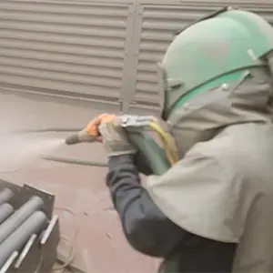 Blaster in a hood and suit sandblasting steel parts in the blast room